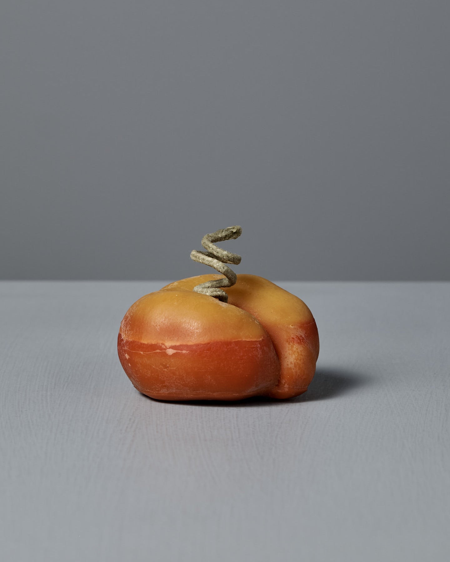 The Flat Peach Candle, designed to resemble a flat peach with a corkscrew detail and beeswax-like finish, sits on a gray surface against a gray background.