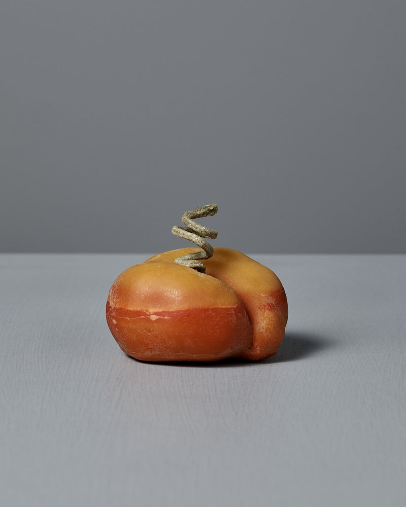 The Flat Peach Candle, designed to resemble a flat peach with a corkscrew detail and beeswax-like finish, sits on a gray surface against a gray background.