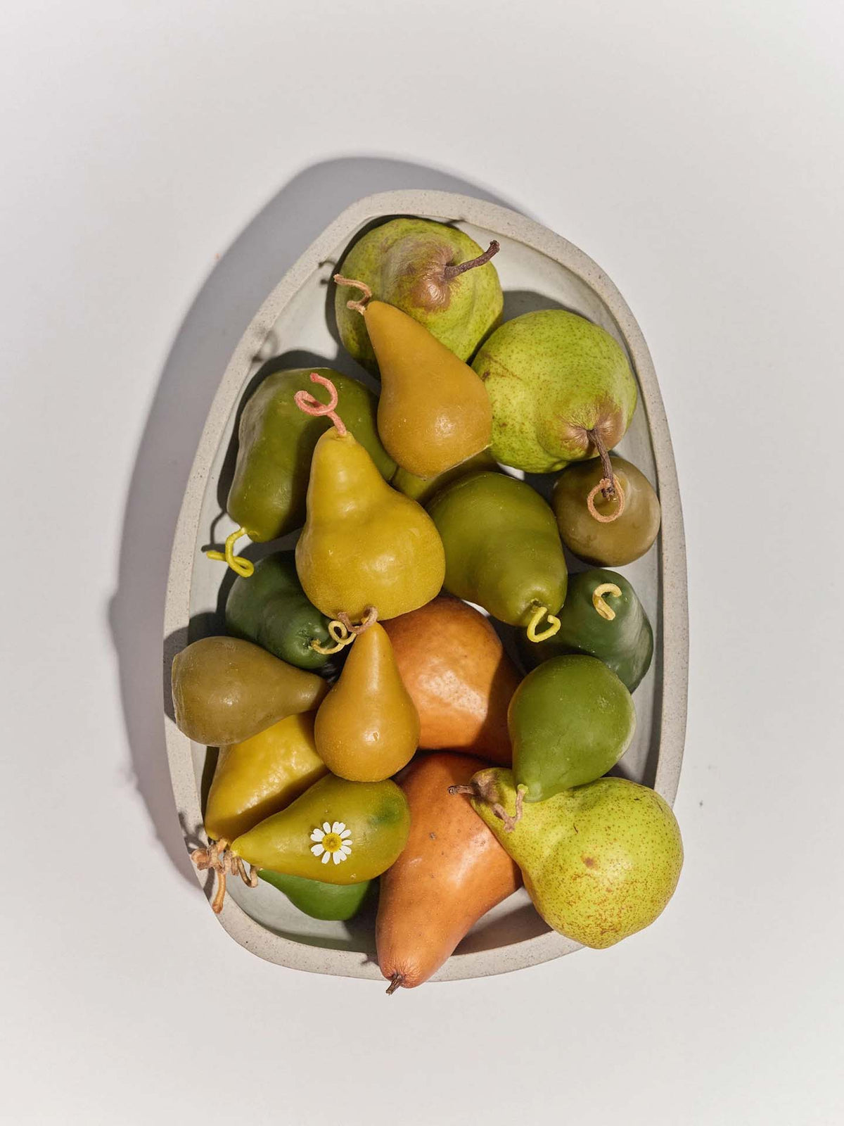 A white oval dish with assorted yellow, green, and orange pears is displayed beside a Green Pear Candle – Large, creating a cozy arrangement.