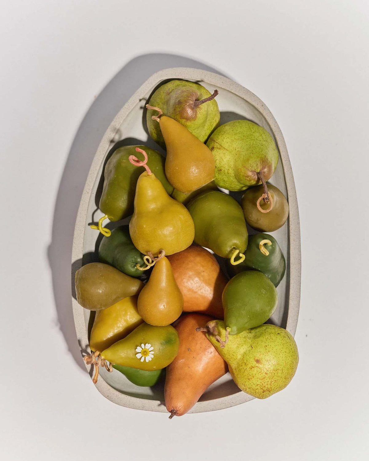 A white oval dish with assorted yellow, green, and orange pears is displayed beside a Green Pear Candle – Large, creating a cozy arrangement.