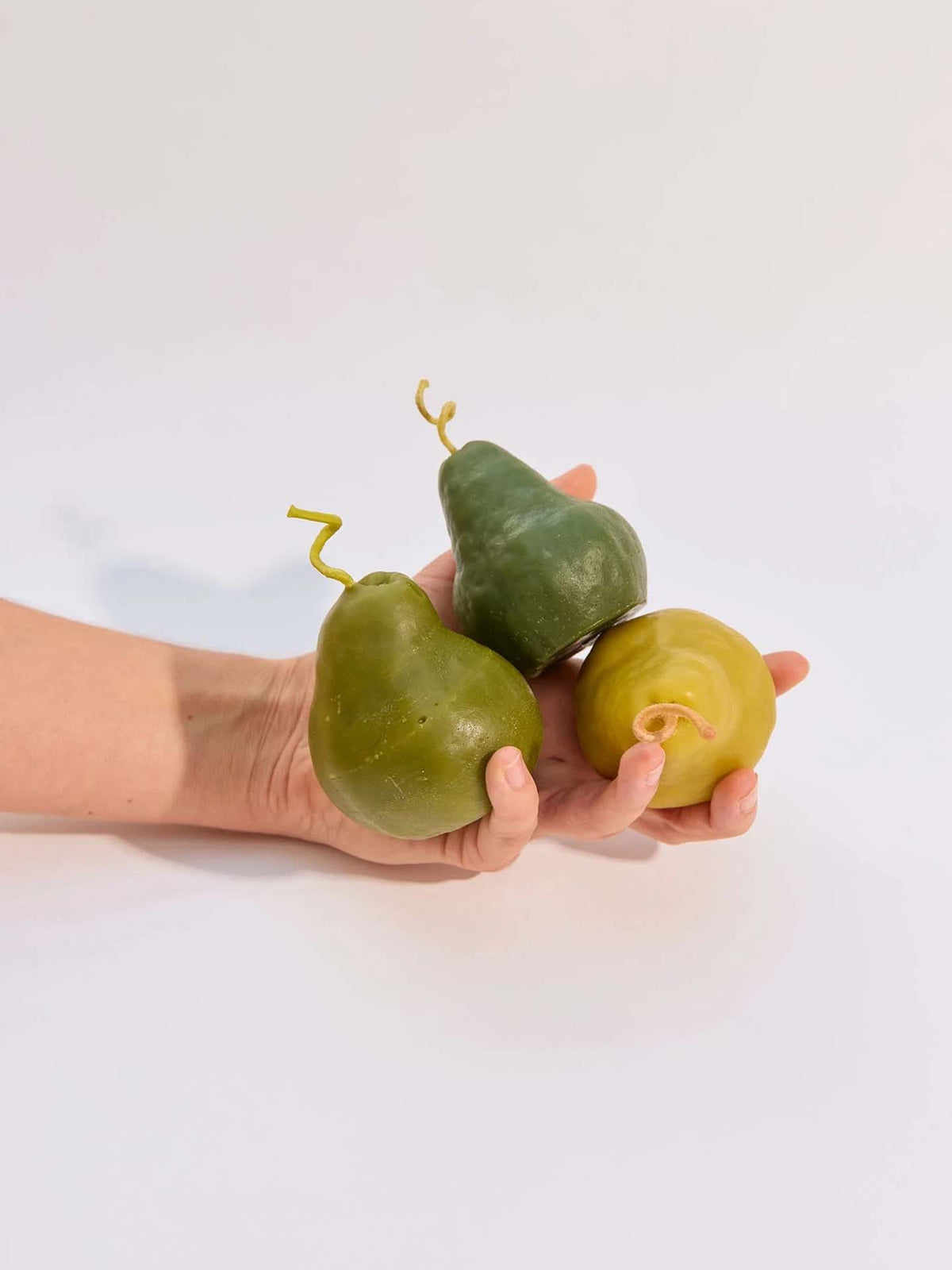 A hand holds two Green Pear Candle – Large and one round yellow beeswax candle against a plain white background.