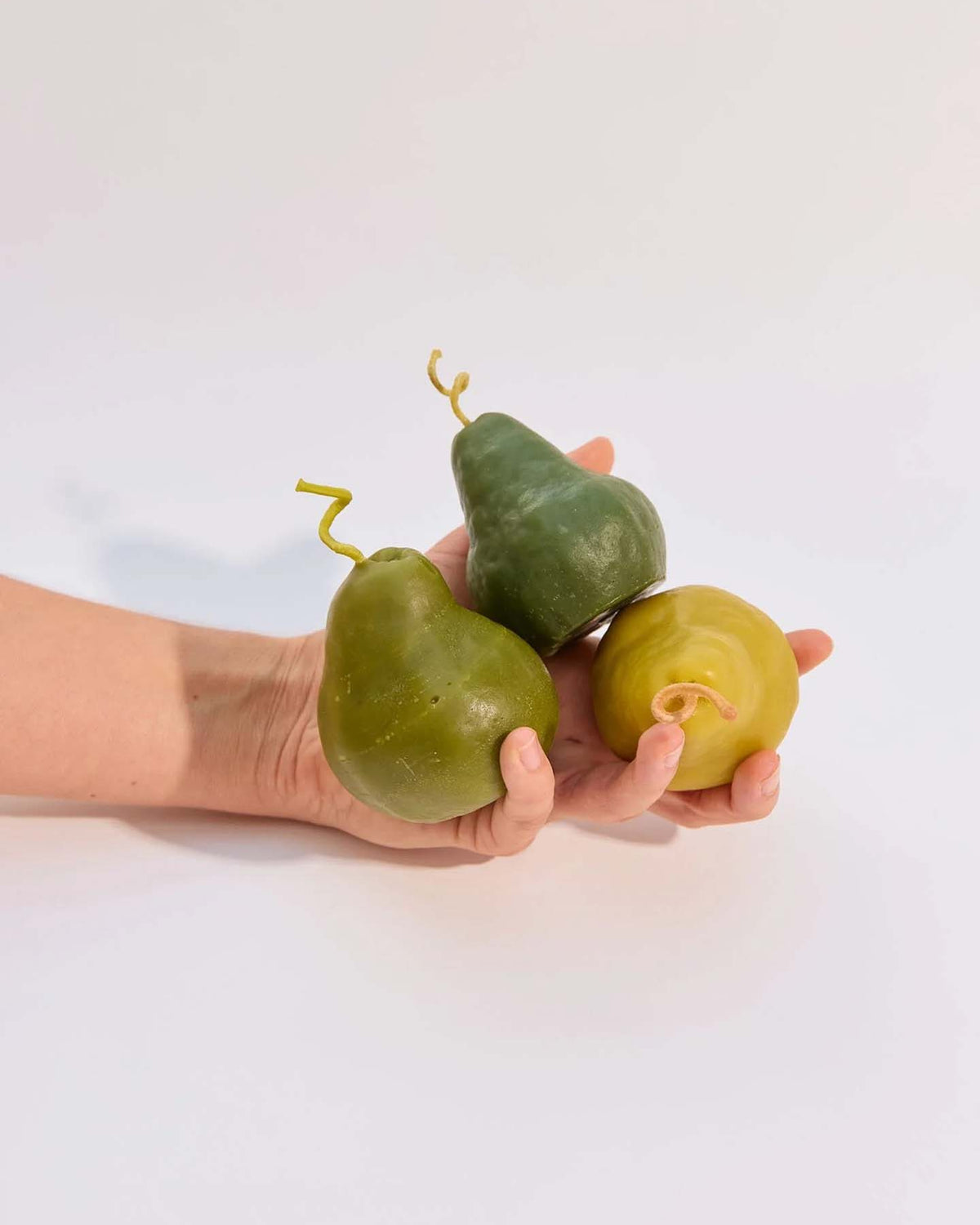 A hand holds two Green Pear Candle – Large and one round yellow beeswax candle against a plain white background.