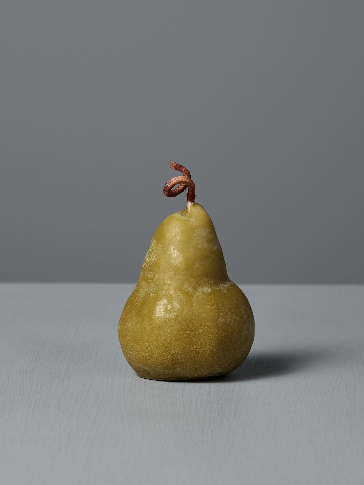 A large green pear-shaped beeswax candle with a twisted wick sits on a light gray surface against a plain gray background.
