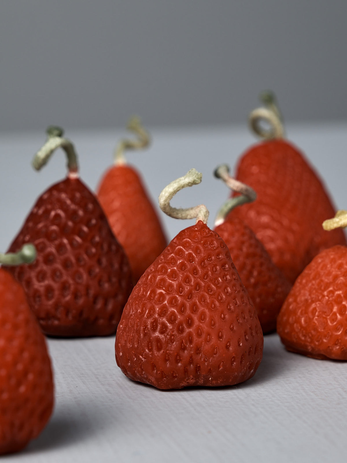 A box of 10 charming handmade Strawberry Candles, designed to look like red strawberries with curly stems, is arranged on a light gray surface against a plain background.