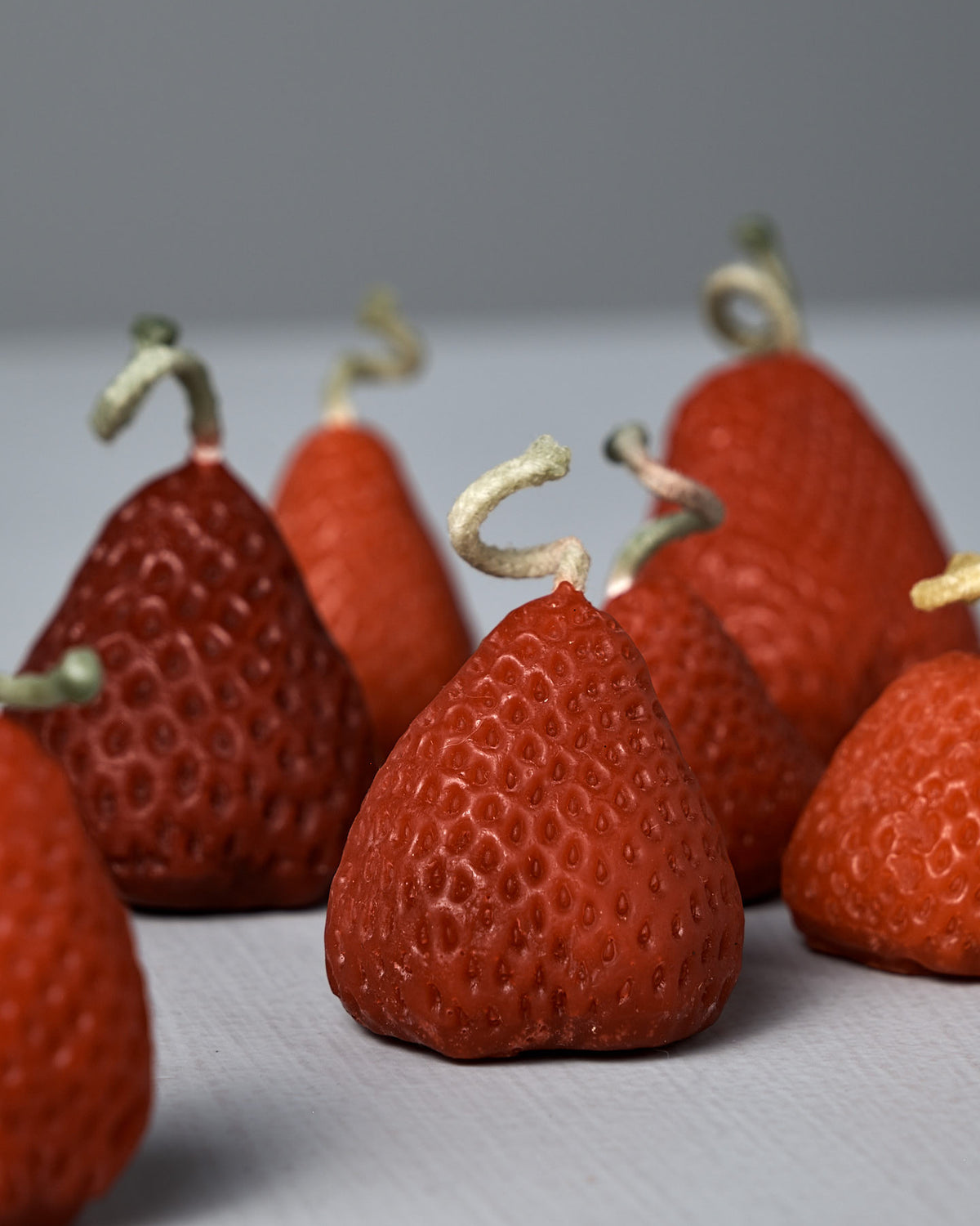 A box of 10 charming handmade Strawberry Candles, designed to look like red strawberries with curly stems, is arranged on a light gray surface against a plain background.