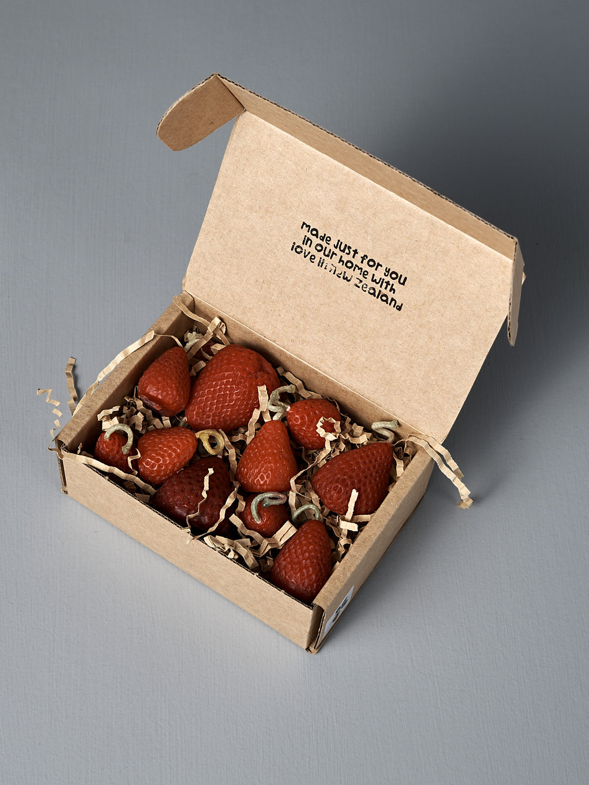 A box of 10 handmade Strawberry Candles rests on shredded paper inside an open cardboard box, with a printed message displayed on the inner lid.
