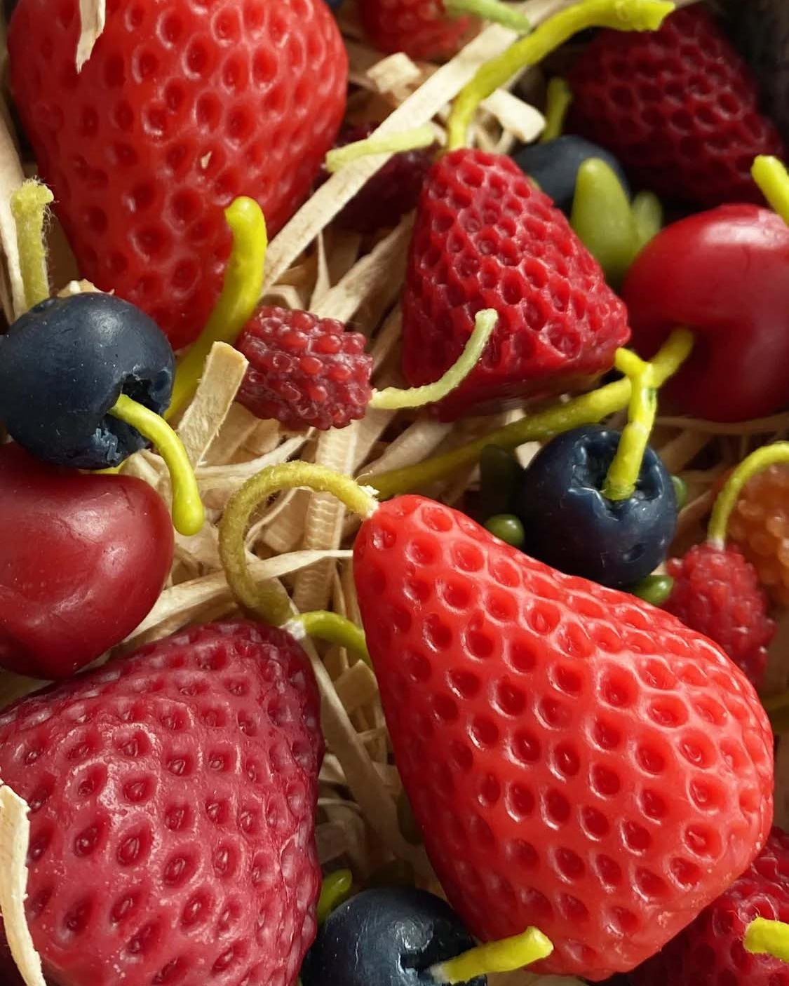 A close-up of Strawberry Candles—Box of 10, shaped like strawberries and nestled in straw-like material beside handmade pure New Zealand beeswax candles.