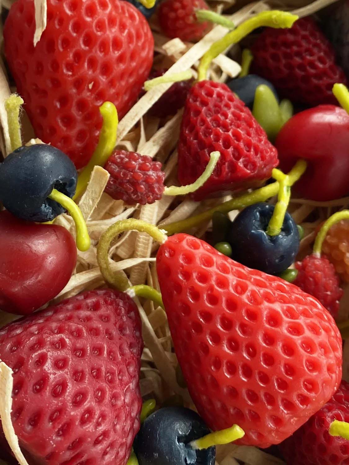 A close-up of Strawberry Candles—Box of 10, shaped like strawberries and nestled in straw-like material beside handmade pure New Zealand beeswax candles.