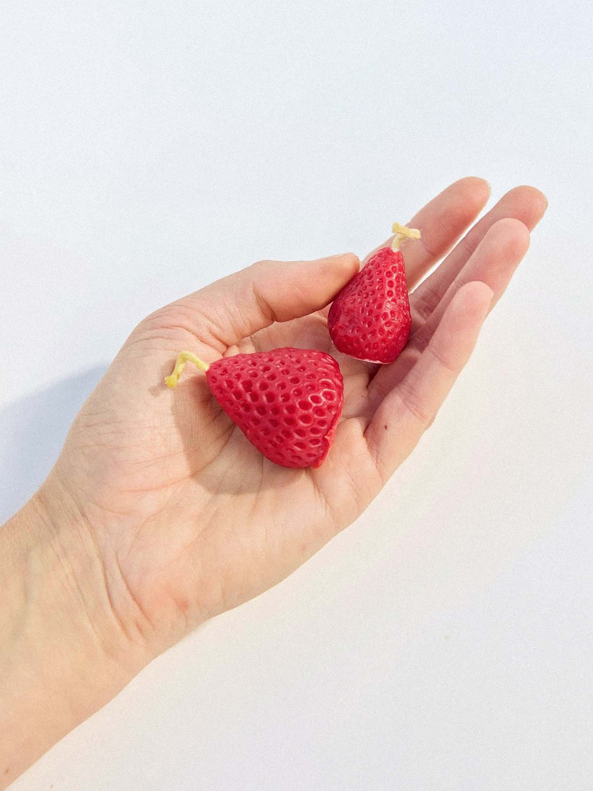 A hand holds two red, strawberry-shaped candles with yellow string at the tops from the Strawberry Candles – Box of 10 set, designed to look like strawberries and crafted from pure New Zealand beeswax.