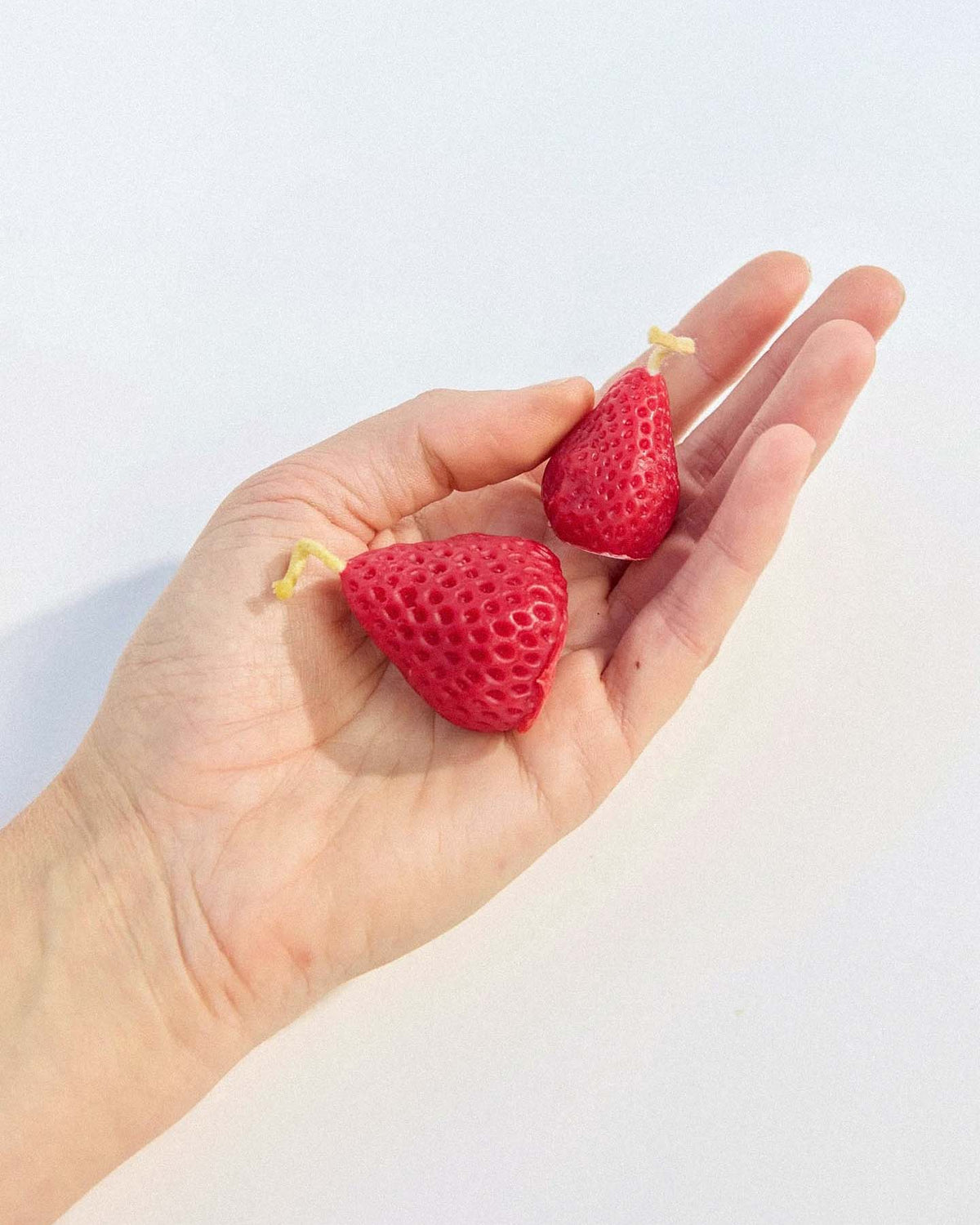 A hand holds two red, strawberry-shaped candles with yellow string at the tops from the Strawberry Candles – Box of 10 set, designed to look like strawberries and crafted from pure New Zealand beeswax.