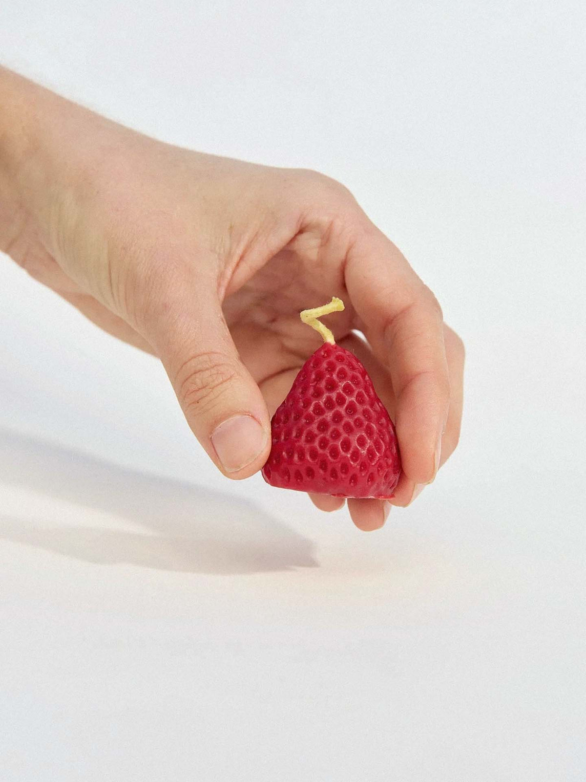 A hand holds one of the Strawberry Candles – Box of 10, handmade from New Zealand beeswax and shaped like a strawberry with a pear's texture and stem, set against a plain background.