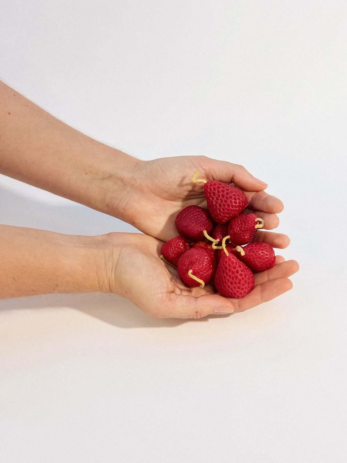 Two hands hold a box of 10 handmade Strawberry Candles, each featuring mini banana peels as stems, against a plain background.