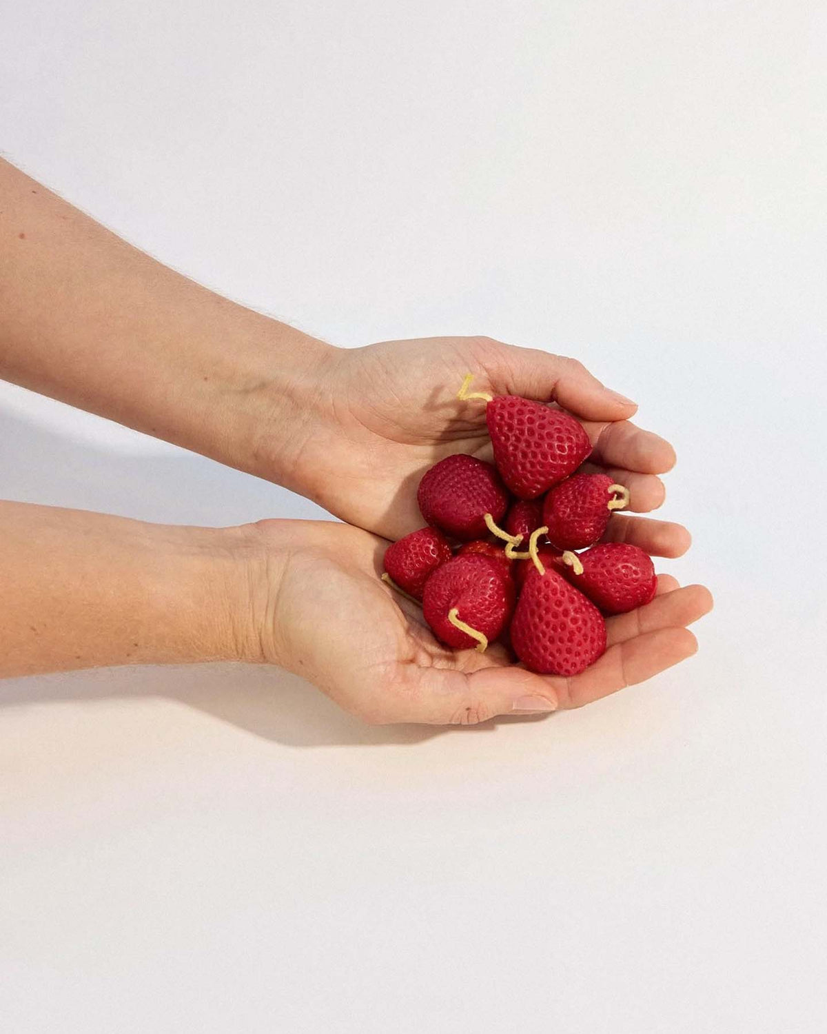 Two hands hold a box of 10 handmade Strawberry Candles, each featuring mini banana peels as stems, against a plain background.