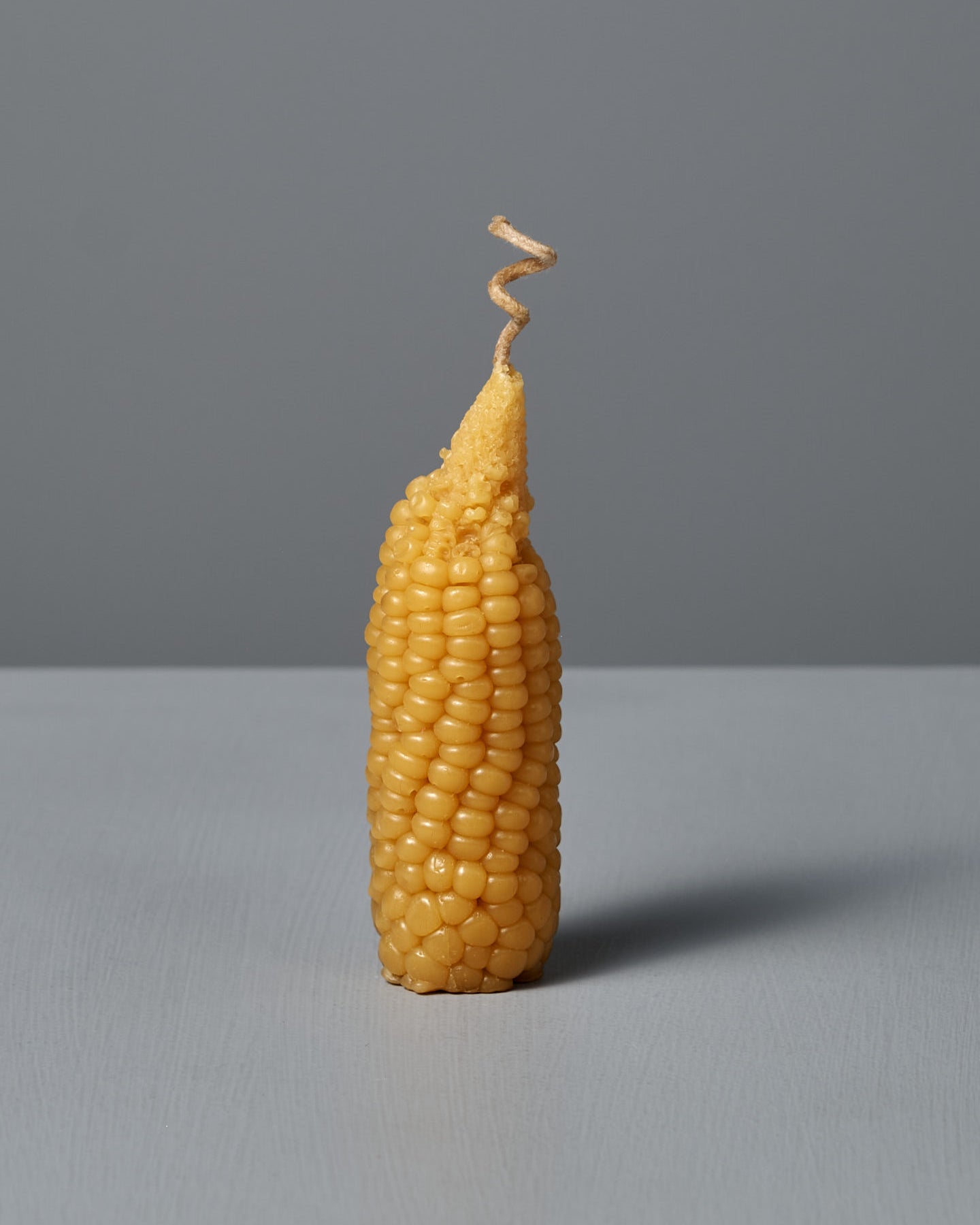 A single Sweetcorn Candle, crafted to mimic an ear of corn with a corn silk twist at the top, stands upright on a flat surface against a plain gray background, resembling real sweetcorn made from pure New Zealand beeswax.