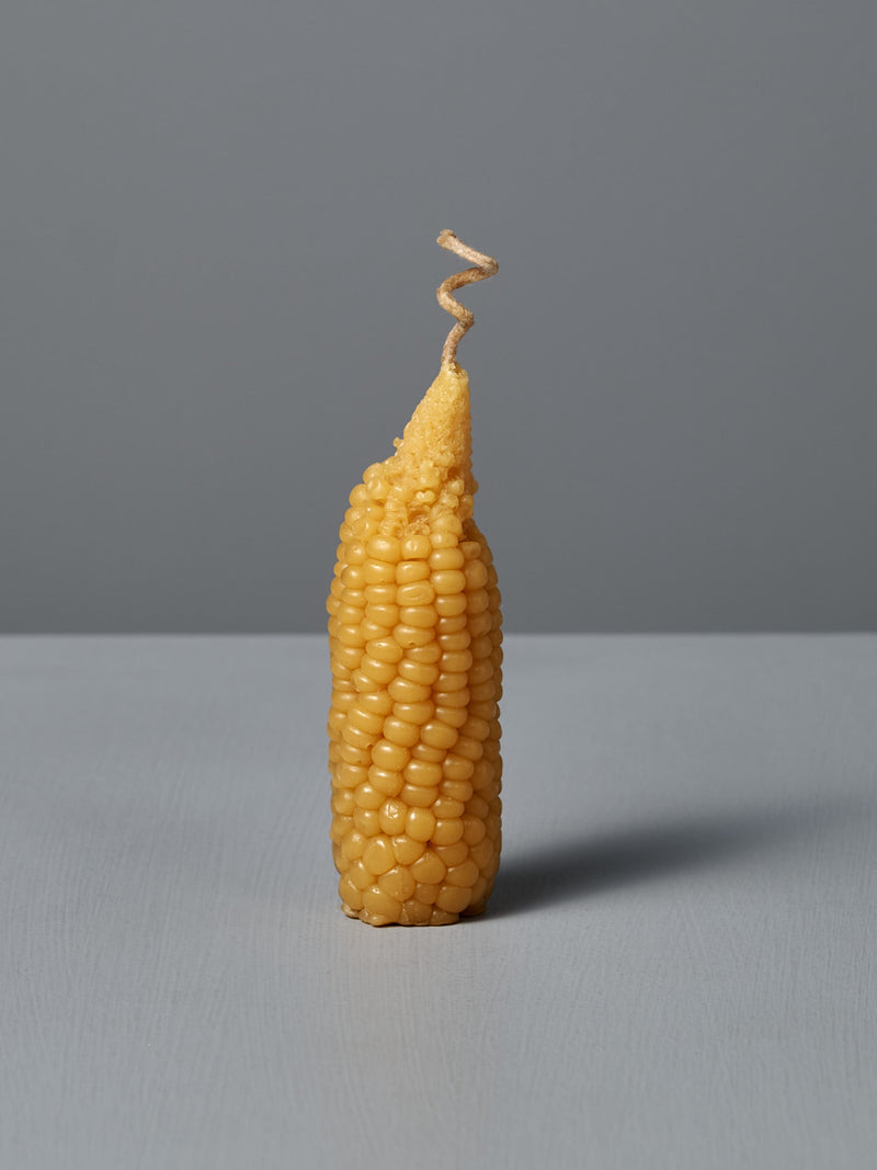 A single Sweetcorn Candle, crafted to mimic an ear of corn with a corn silk twist at the top, stands upright on a flat surface against a plain gray background, resembling real sweetcorn made from pure New Zealand beeswax.