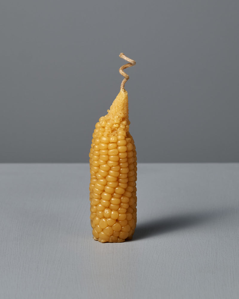 A single Sweetcorn Candle, crafted to mimic an ear of corn with a corn silk twist at the top, stands upright on a flat surface against a plain gray background, resembling real sweetcorn made from pure New Zealand beeswax.