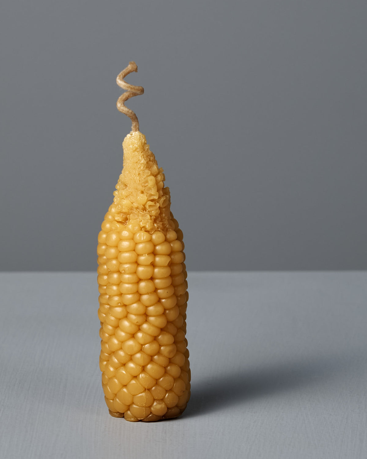 The Sweetcorn Candle, crafted from pure New Zealand beeswax and shaped like an ear of corn, stands upright on a flat gray surface against a plain gray background.