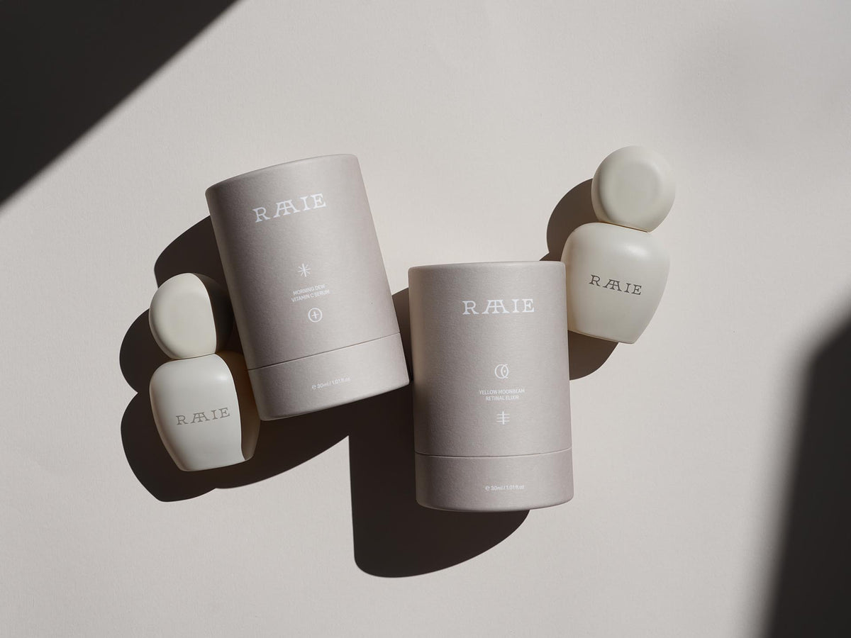 The image displays four RAAIE skincare products, including the AM/ PM Set, consisting of two cylindrical containers and two bottle-shaped items, placed on a neutral-colored surface with shadows.