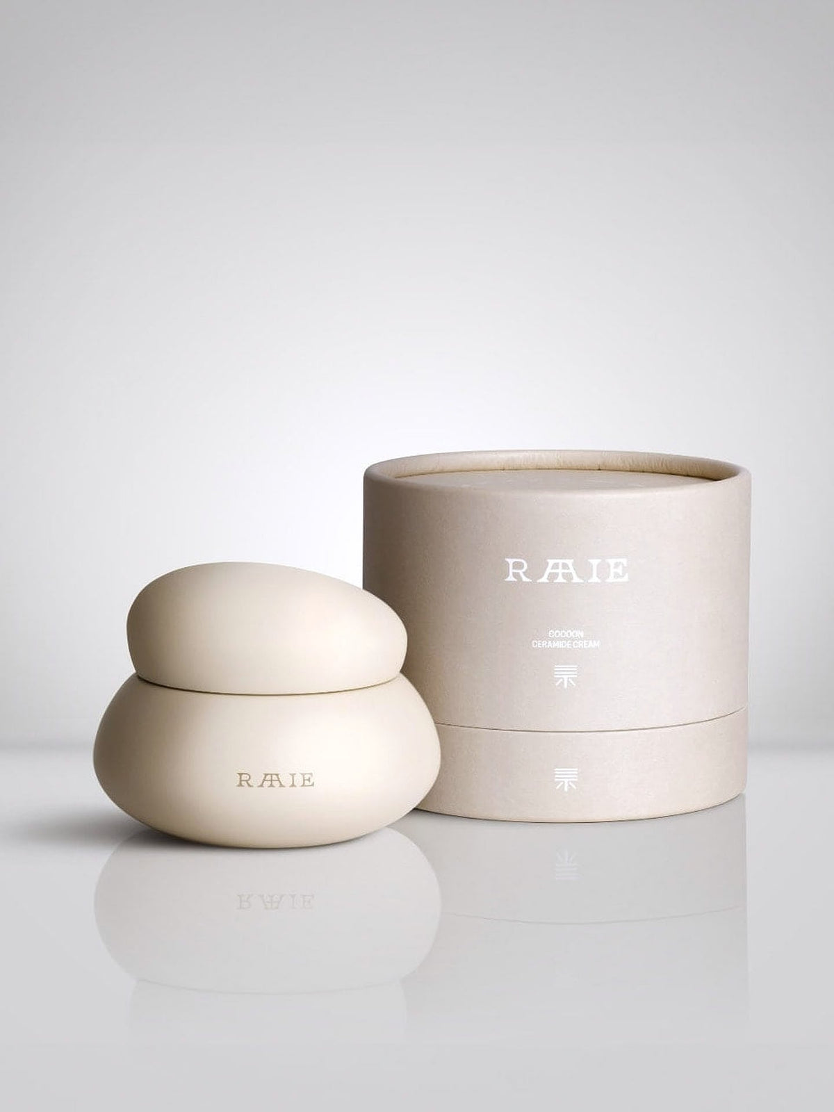 Minimalist cream-colored cosmetics container with &quot;RAAIE&quot; branding, shaped in a rounded form, placed next to its matching cylindrical packaging. Perfect for a hydrating moisturiser like the Cocoon Ceramide Cream that helps rebuild the skin barrier.