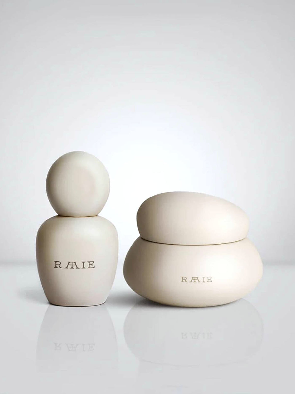 Two sleek, beige skincare containers labeled &quot;RAAIE&quot; are placed side by side against a white background. The left container, holding Lunar Repair Duo, is vertically elongated, while the right one features a rounded, pebble-like shape ideal for Lunar Repair Duo—both promising luminous skin.