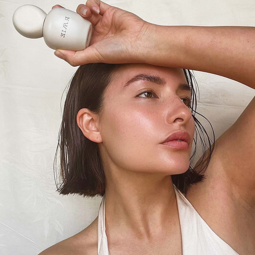 A person with short brown hair holds a white bottle labeled &quot;RAAIE Lunar Repair Duo&quot; against their forehead. They have a neutral expression and are looking to the side.