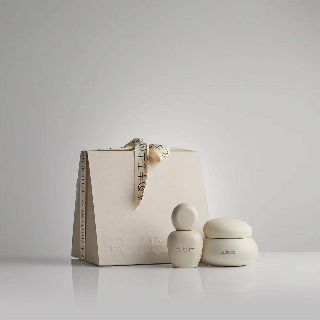 A beige gift bag adorned with a patterned ribbon stands next to two sleek, small ceramic containers labeled &quot;RAAIE&quot; on a plain white background, promising luminous skin with their Lunar Repair Duo.