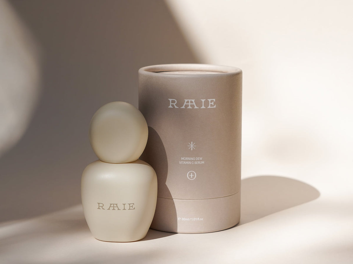 A cream-colored product bottle labeled &quot;RAAIE&quot; next to a matching cylindrical container on a minimalistic beige background, featuring Morning Dew Vitamin C Serum enriched with New Zealand botanicals.