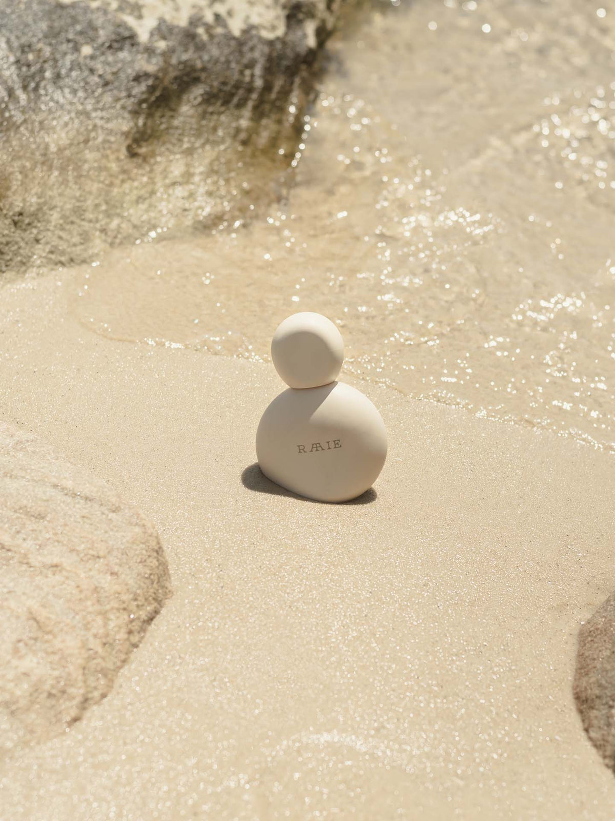 A small, round white bottle with the word &quot;RAAIE&quot; on it sits on wet sand near ocean water and rocks, offering broad spectrum protection with its lightweight formula.