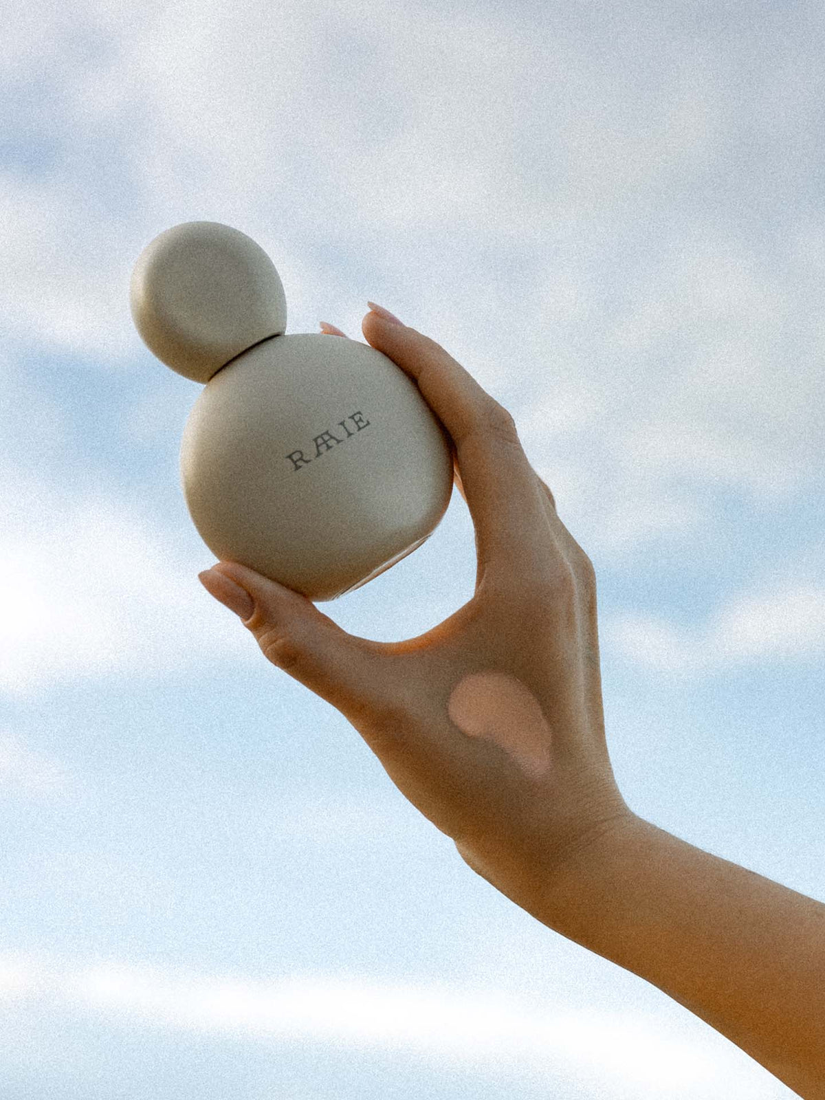 A hand holds a round, light-colored bottle labeled &quot;RAAIE Sun Milk Drops Tinted SPF 50+&quot; against a backdrop of a cloudy sky, showcasing its lightweight formula and broad spectrum protection.