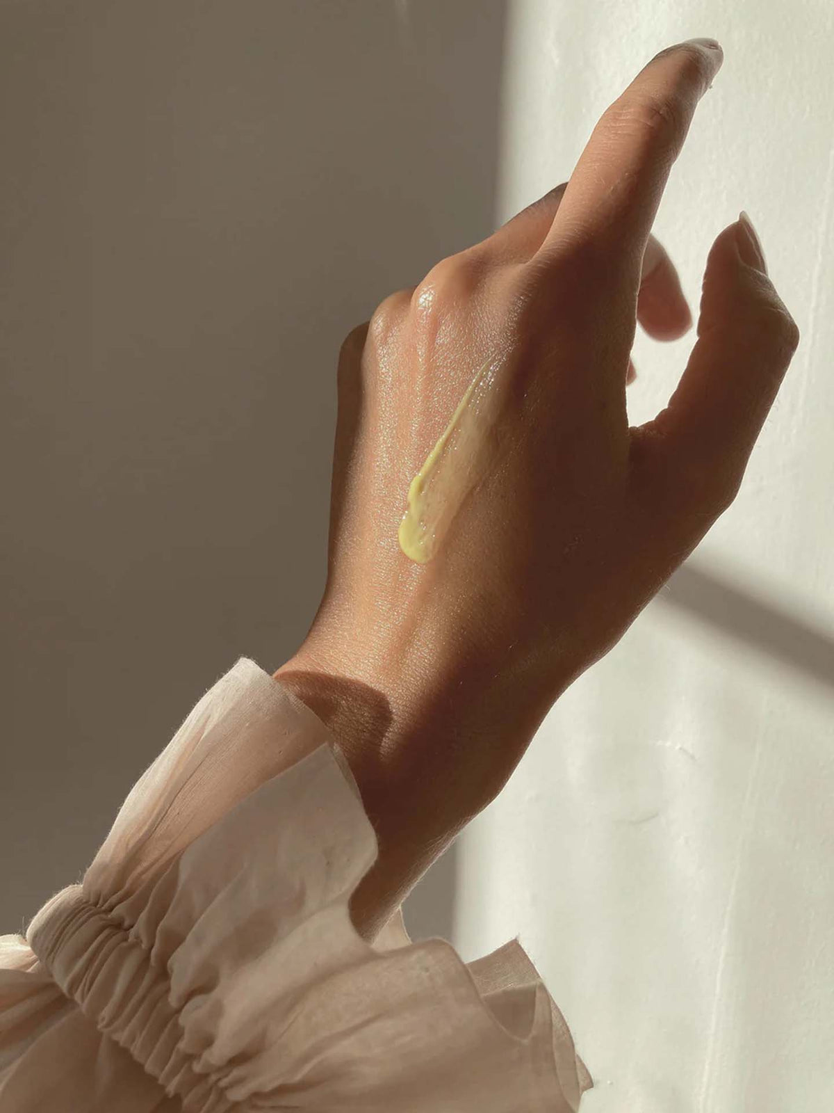 A hand with a streak of RAAIE Yellow Moonbeam Retinal Elixir on the back, partially lit by sunlight. The hand is wearing a sheer, ruffled sleeve.