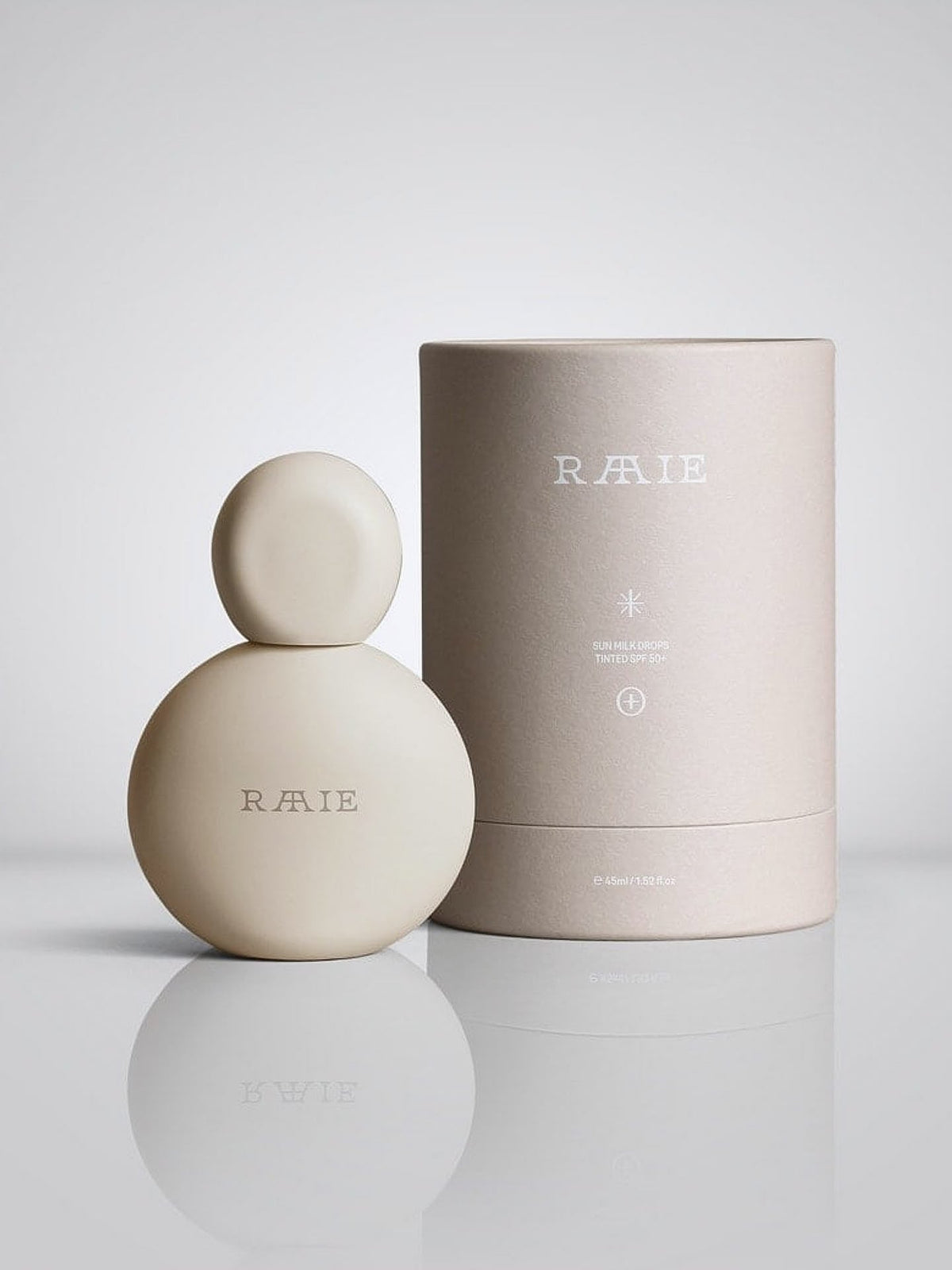 A round, minimalist perfume bottle with &quot;RAAIE&quot; branding sits elegantly next to a cylindrical beige packaging container on a reflective surface, evoking the same sophistication as an ultra-lightweight Sun Milk Drops Tinted SPF 50+.