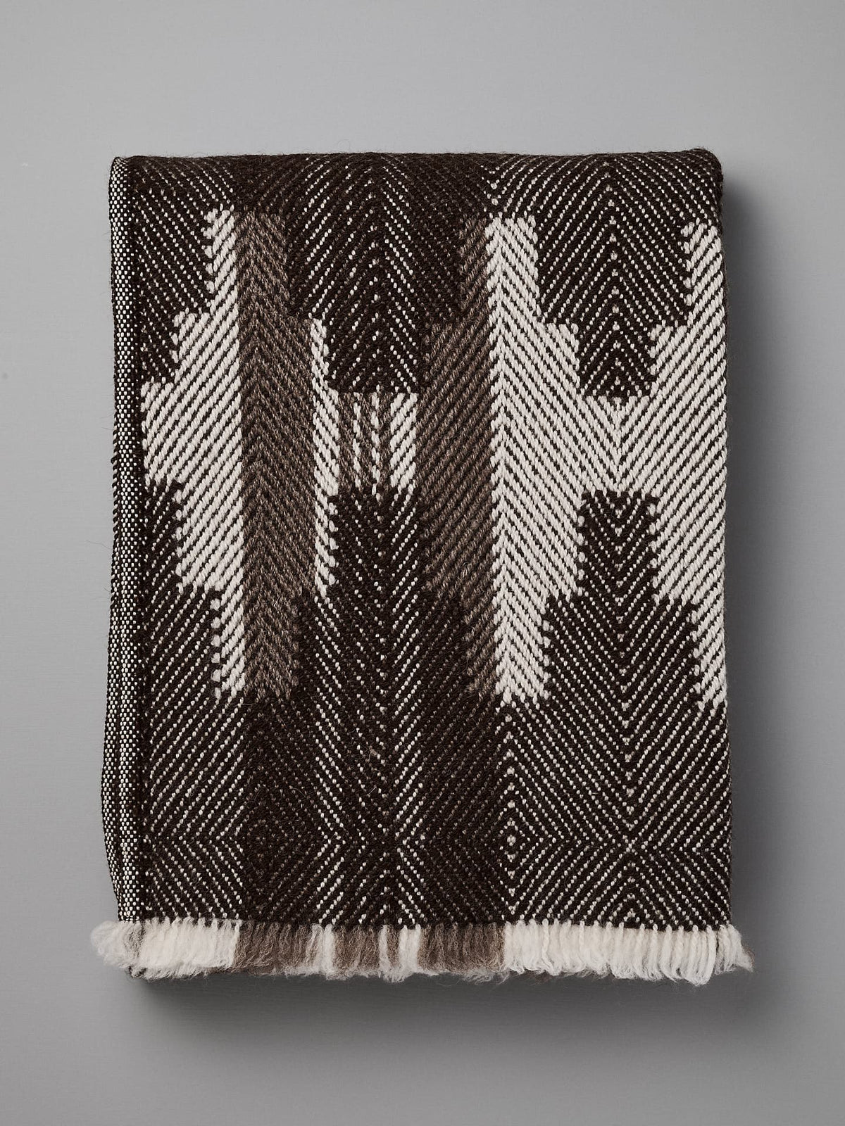 A folded, black and white patterned blanket with geometric designs, reminiscent of traditional Bulgarian shevitza, is displayed against a plain gray background. 

Replace with: 

A folded Rodopska Takan &quot;Bulgarian Shevitza Wool Blanket – Brown&quot; with geometric designs, reminiscent of traditional Bulgarian shevitza, is displayed against a plain gray background.