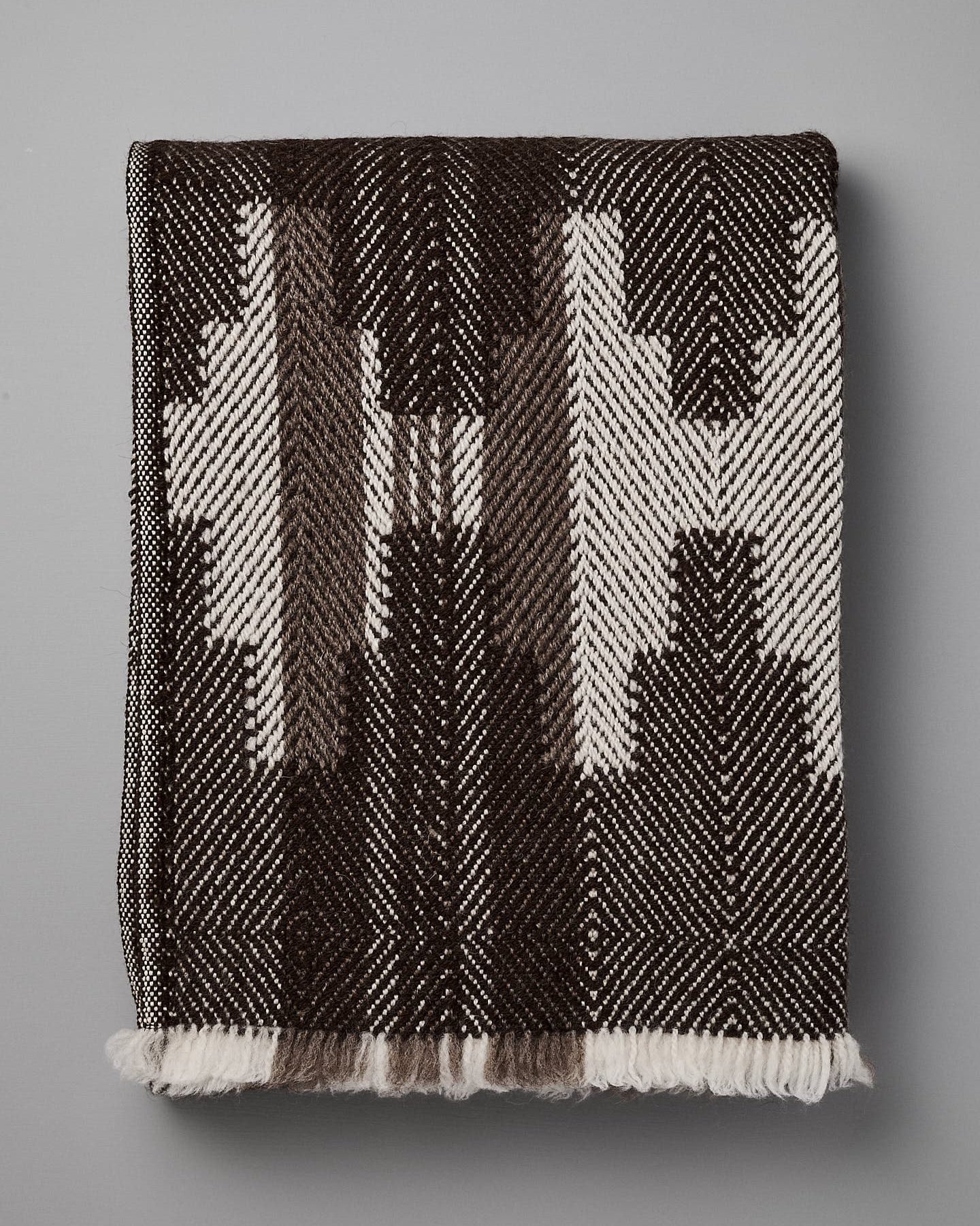 A folded, black and white patterned blanket with geometric designs, reminiscent of traditional Bulgarian shevitza, is displayed against a plain gray background. 

Replace with: 

A folded Rodopska Takan "Bulgarian Shevitza Wool Blanket – Brown" with geometric designs, reminiscent of traditional Bulgarian shevitza, is displayed against a plain gray background.