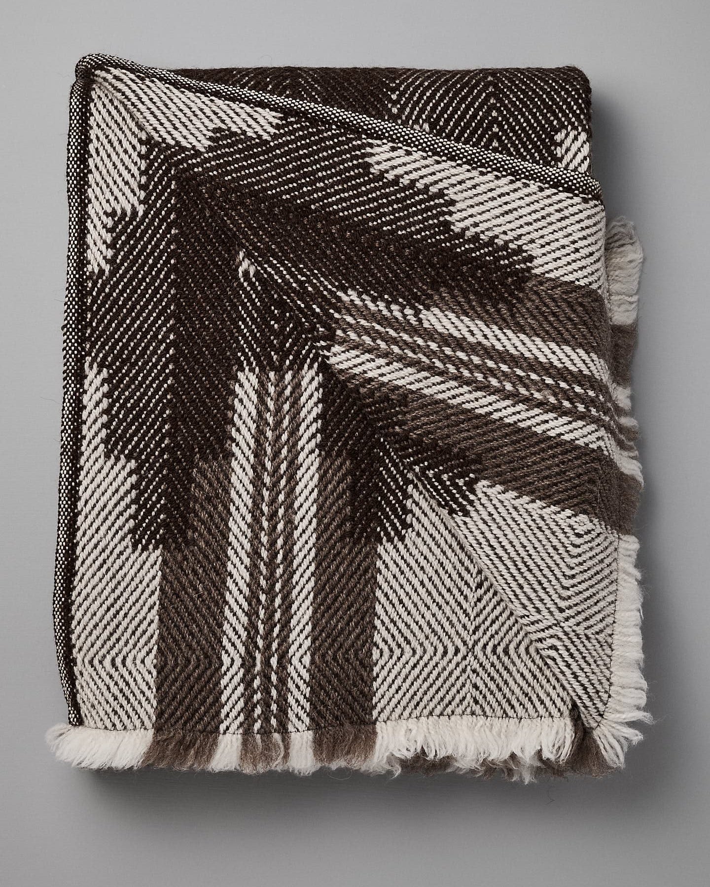 A folded, black and white patterned blanket with geometric designs, reminiscent of traditional Bulgarian shevitza, is displayed against a plain gray background. 

Replace with: 

A folded Rodopska Takan "Bulgarian Shevitza Wool Blanket – Brown" with geometric designs, reminiscent of traditional Bulgarian shevitza, is displayed against a plain gray background.