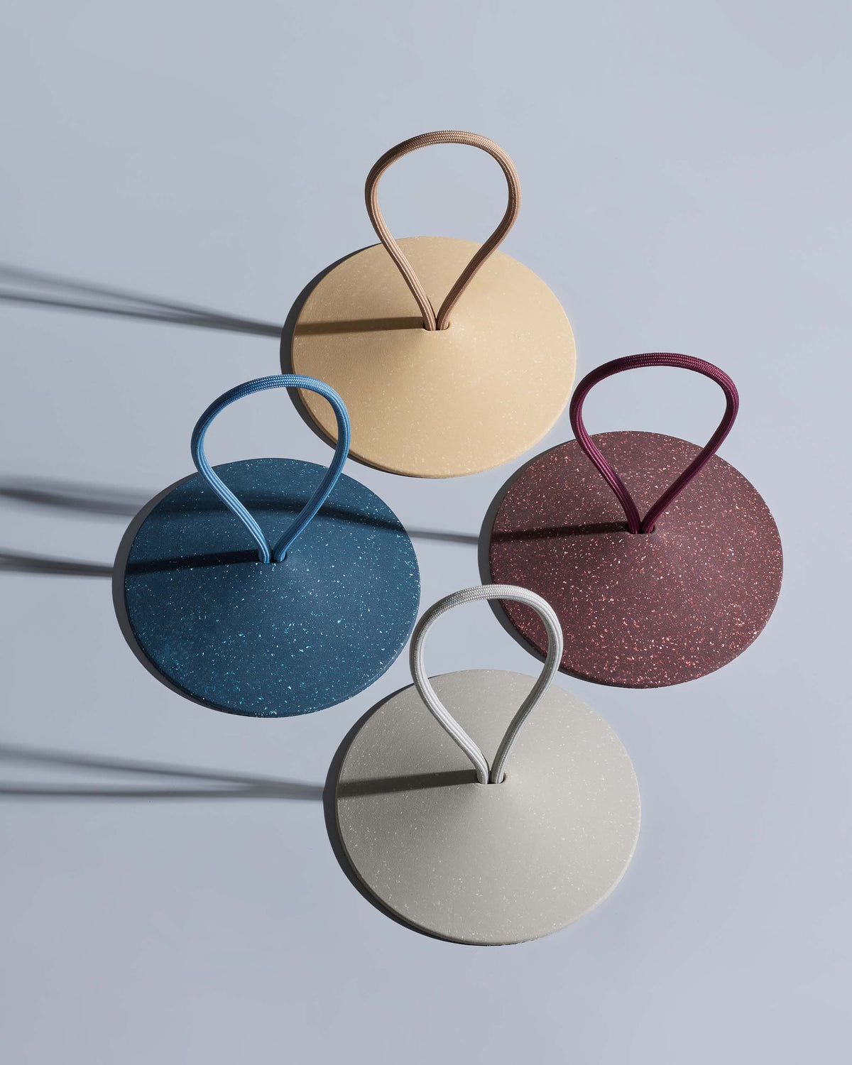 Four round, flat objects in beige, blue, burgundy, and light gray with looped handles, arranged in a square pattern on a light surface casting long shadows.