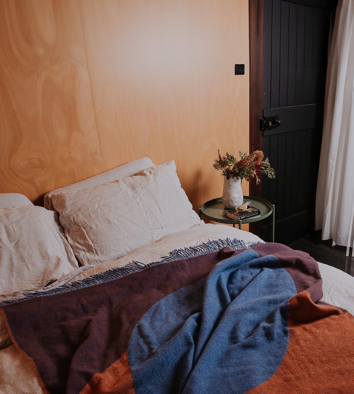 A neatly made bed with white pillows and a colorful blanket. A small side table beside the bed holds a vase of flowers and a pair of glasses. The room features a wooden wall and a dark door.