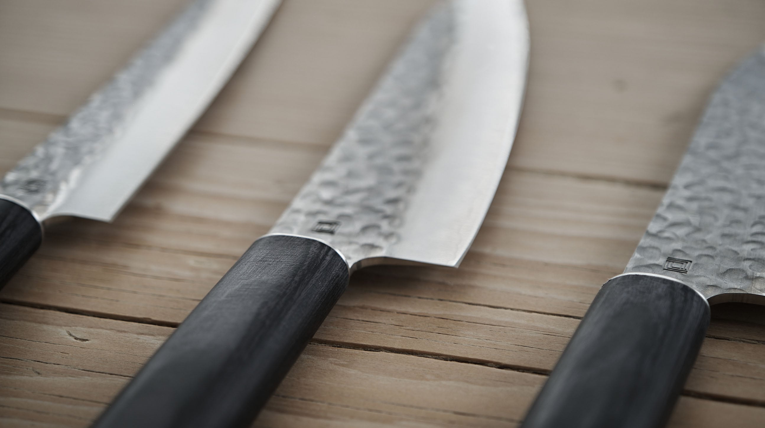 Close-up of three chef knives with hammered blades and dark wooden handles arranged on a wooden surface.