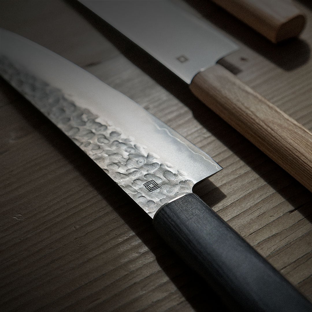 Close-up of two Japanese chef knives with wooden handles displayed on a wooden surface. One knife features a hammered texture on the blade.