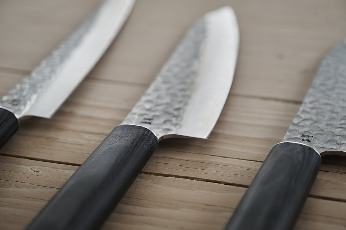 Close-up view of three Yamato Yanagiba knives with hammered Sanmai steel blades and dark handles by Shizu Hamono Co., arranged diagonally on a wooden surface, reflecting exquisite Japanese design.