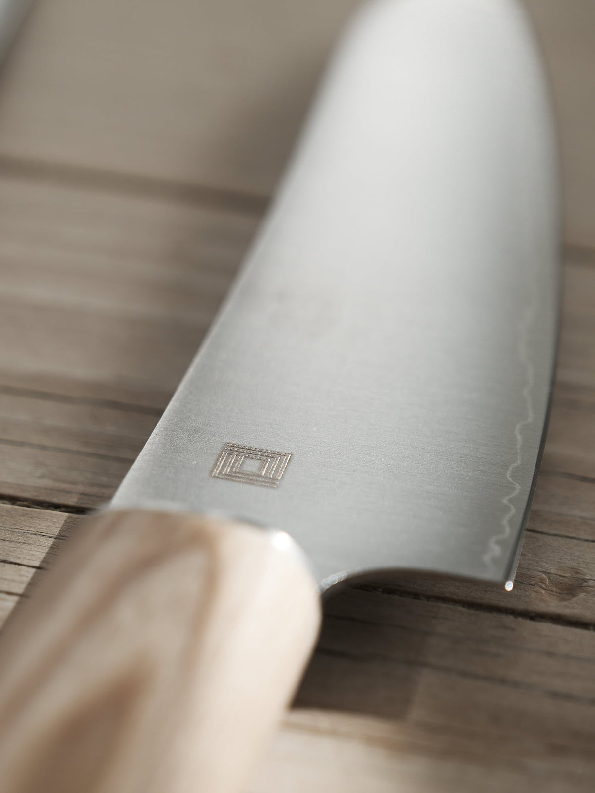 Close-up of a Yuri Santoku Knife by Shizu Hamono Co., featuring a light wooden handle, resting on a wooden surface. The blade showcases an engraved logo and has a sharp edge, ensuring precision and excellence among Japanese chef knives.