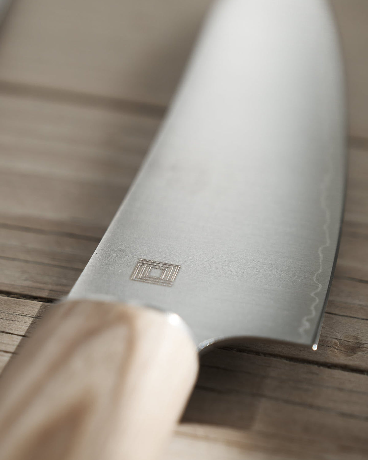 Close-up of a Yuri Santoku Knife by Shizu Hamono Co., featuring a light wooden handle, resting on a wooden surface. The blade showcases an engraved logo and has a sharp edge, ensuring precision and excellence among Japanese chef knives.