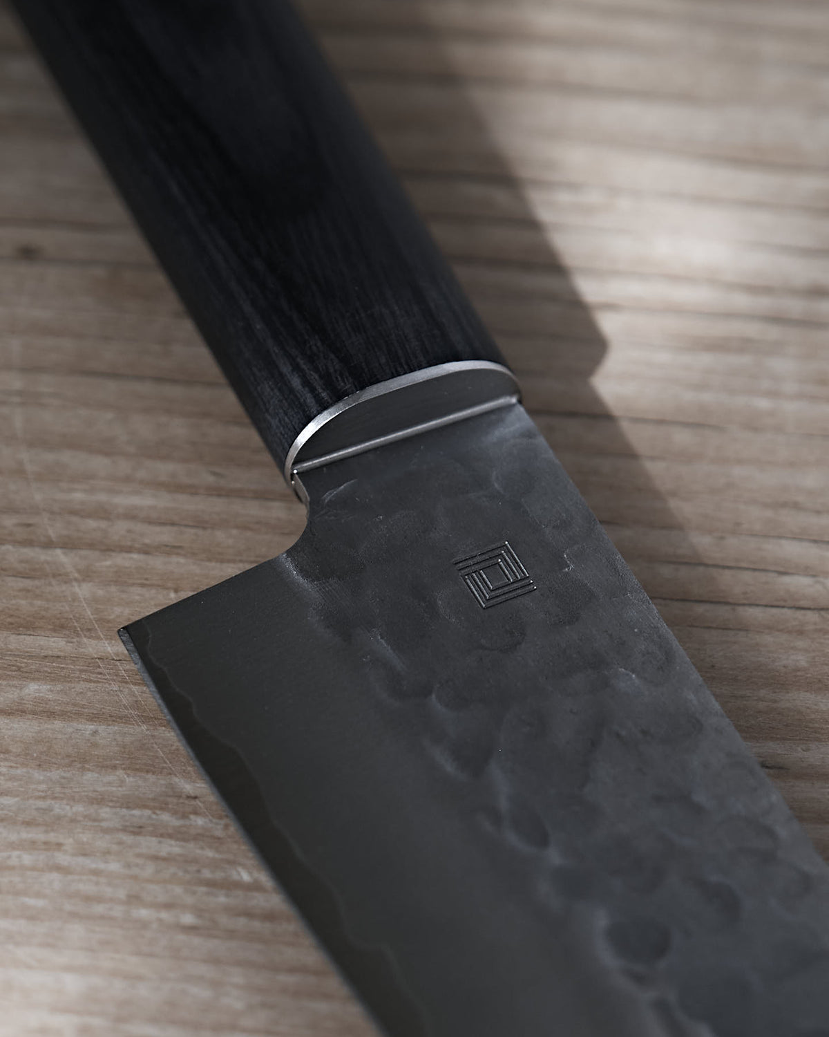 Close-up of a Shizu Hamono Co. Yamato Santoku Knife, featuring a textured 3 Layer Sanmai Steel blade, black handle, and metal bolster, placed on a wooden surface.