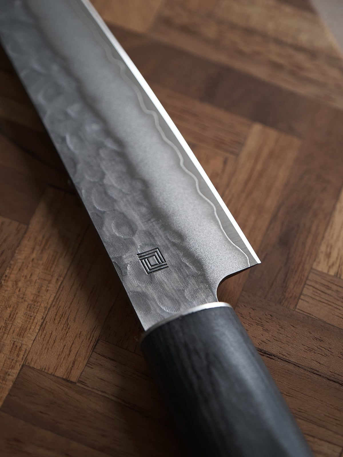 Close-up of a Yamato Yanagiba Knife by Shizu Hamono Co., showcasing its patterned Sanmai steel blade and dark handle, resting on a wooden surface with a herringbone pattern.