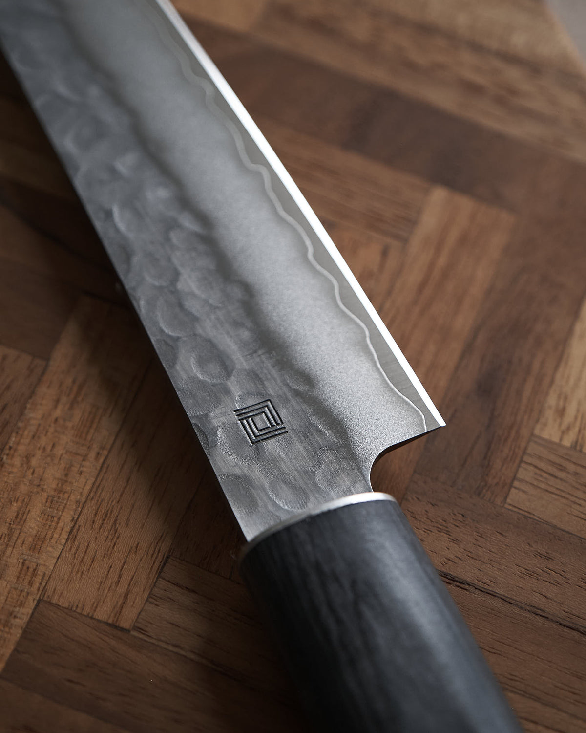 Close-up of a Yamato Yanagiba Knife by Shizu Hamono Co., showcasing its patterned Sanmai steel blade and dark handle, resting on a wooden surface with a herringbone pattern.