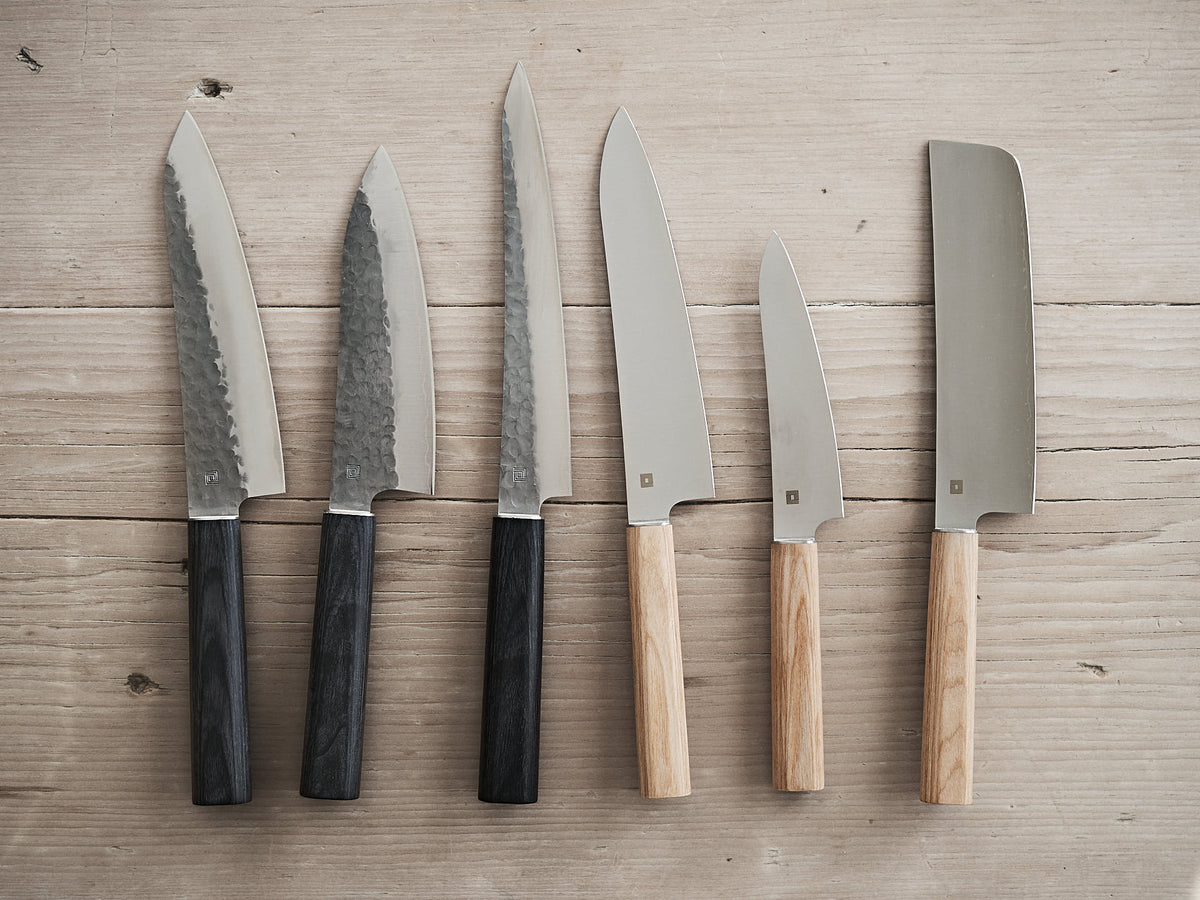 A set of six kitchen knives featuring various blade shapes and sizes, including the Yuri Nakiri Knife by Shizu Hamono Co., perfect for dicing vegetables, with handles made from contrasting dark and light wood, displayed on a wooden surface.