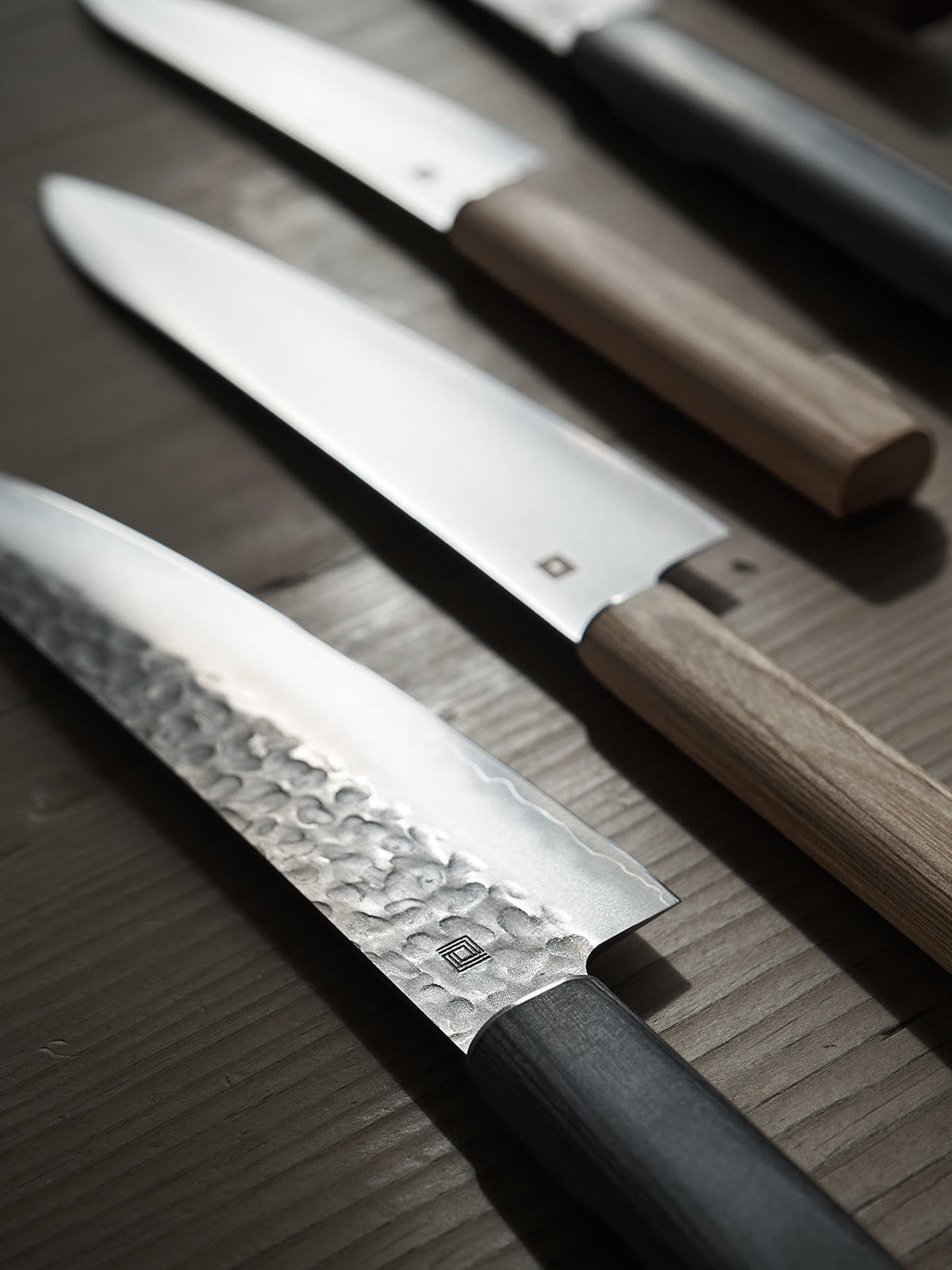 Close-up of four kitchen knives, including a Japanese Chef knife and a Shizu Hamono Co. Yamato Santoku Knife with a 3 Layer Sanmai Steel Blade, each boasting distinct handle materials and blade styles, arranged in a diagonal line on a wooden surface.