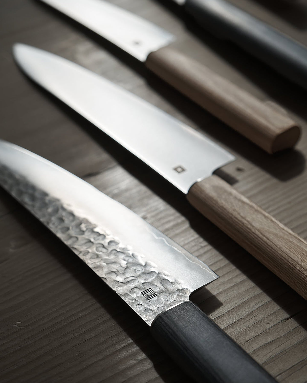 Close-up of four kitchen knives, including a Japanese Chef knife and a Shizu Hamono Co. Yamato Santoku Knife with a 3 Layer Sanmai Steel Blade, each boasting distinct handle materials and blade styles, arranged in a diagonal line on a wooden surface.
