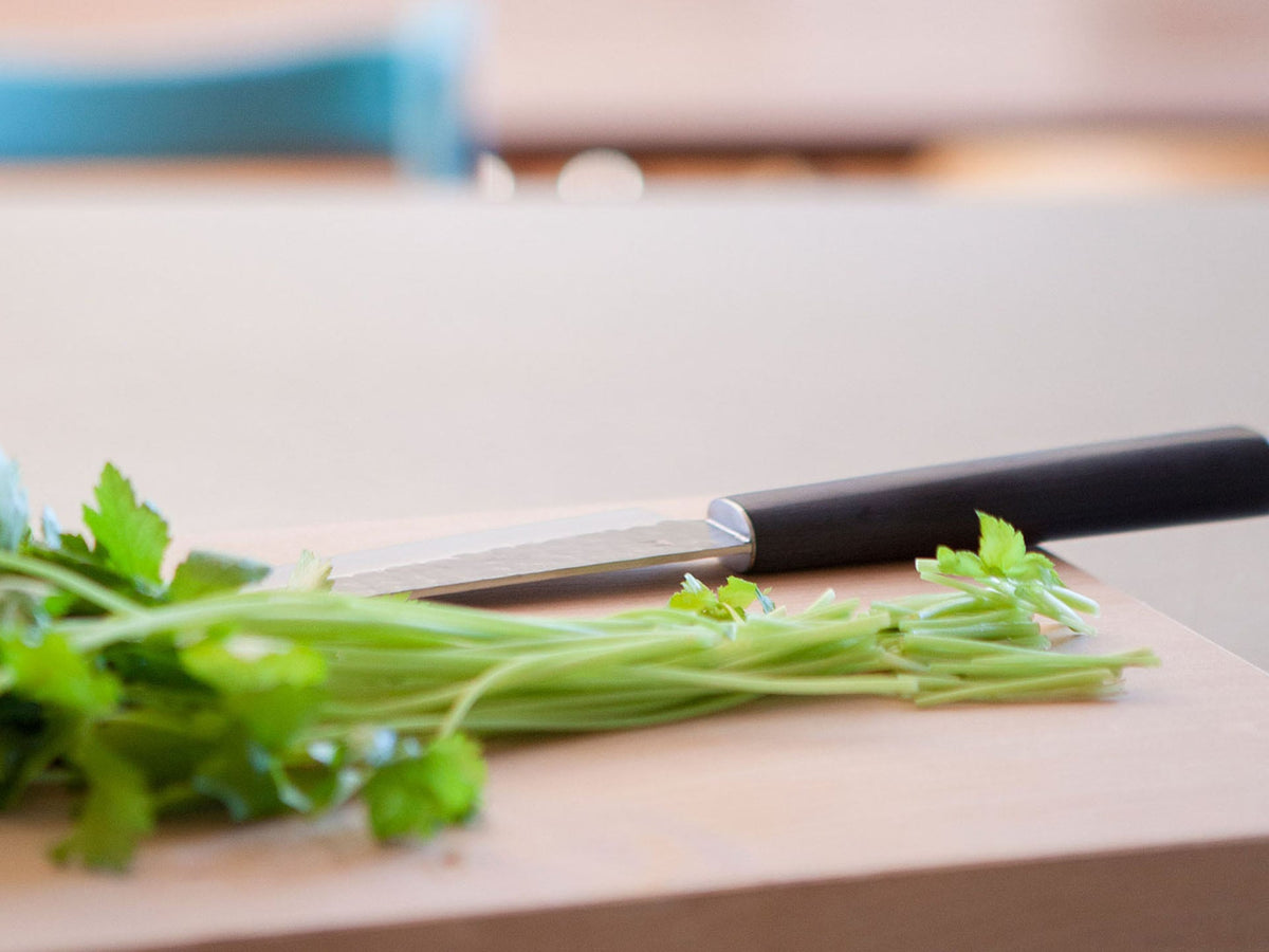 A Yamato Santoku Knife from Shizu Hamono Co., crafted from AUS8 Japanese stainless steel, lies on a wooden cutting board next to a bunch of fresh cilantro.