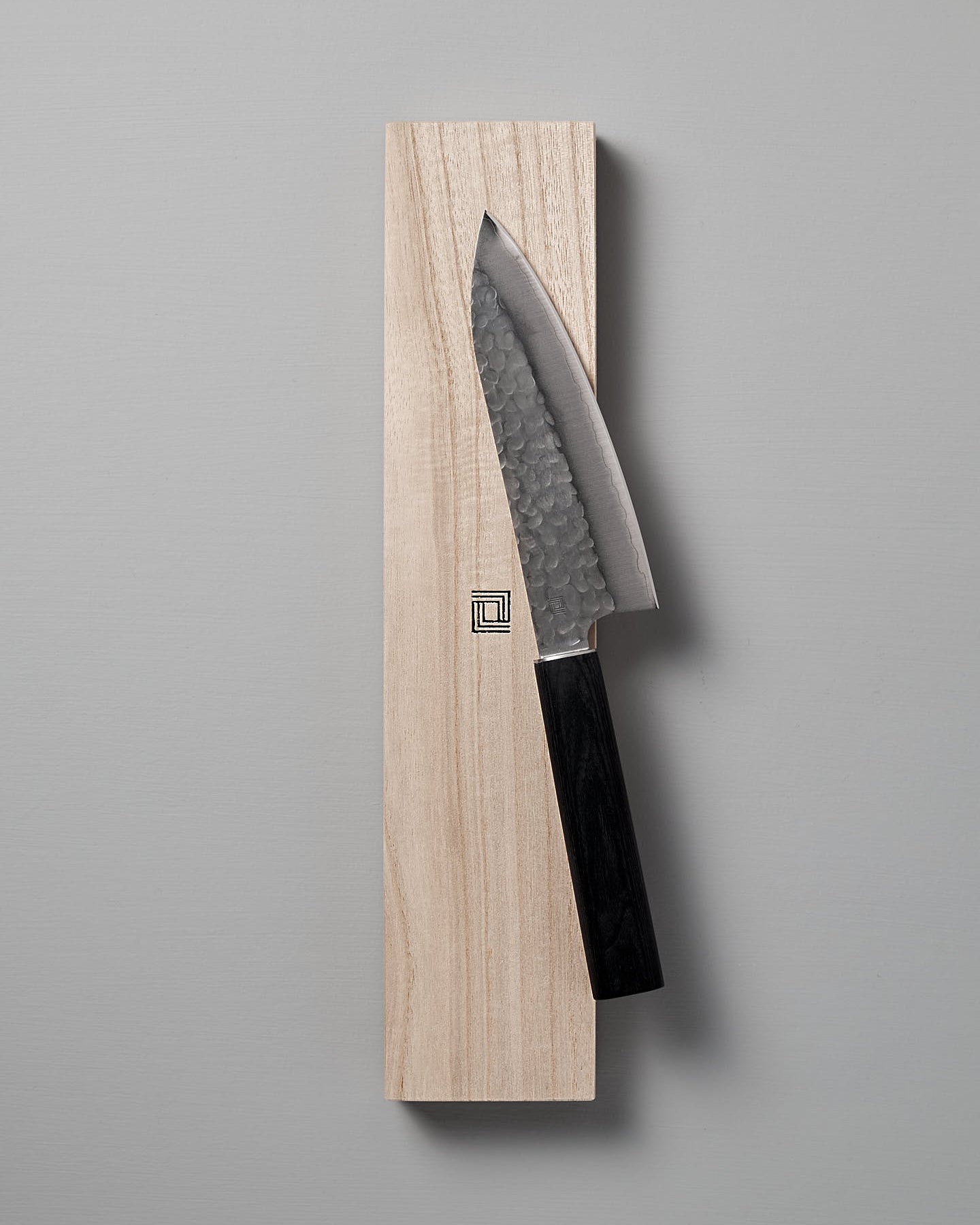 The Yamato Deba Knife by Shizu Hamono Co., featuring a Sanmai steel blade with a hammered finish, rests on a wooden block against a gray background.