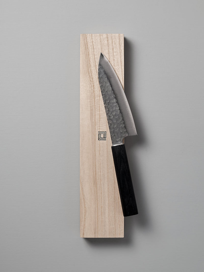 The Yamato Deba Knife by Shizu Hamono Co., featuring a Sanmai steel blade with a hammered finish, rests on a wooden block against a gray background.